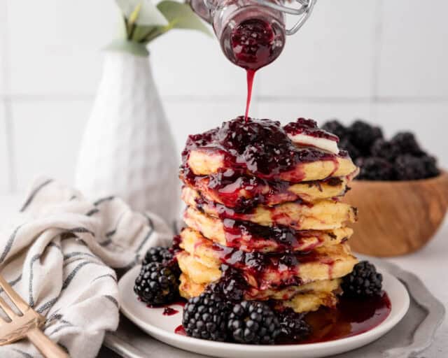 blackberry syrup being drizzled onto a stack of pancakes from the side with more fresh blackberries on the plate and background