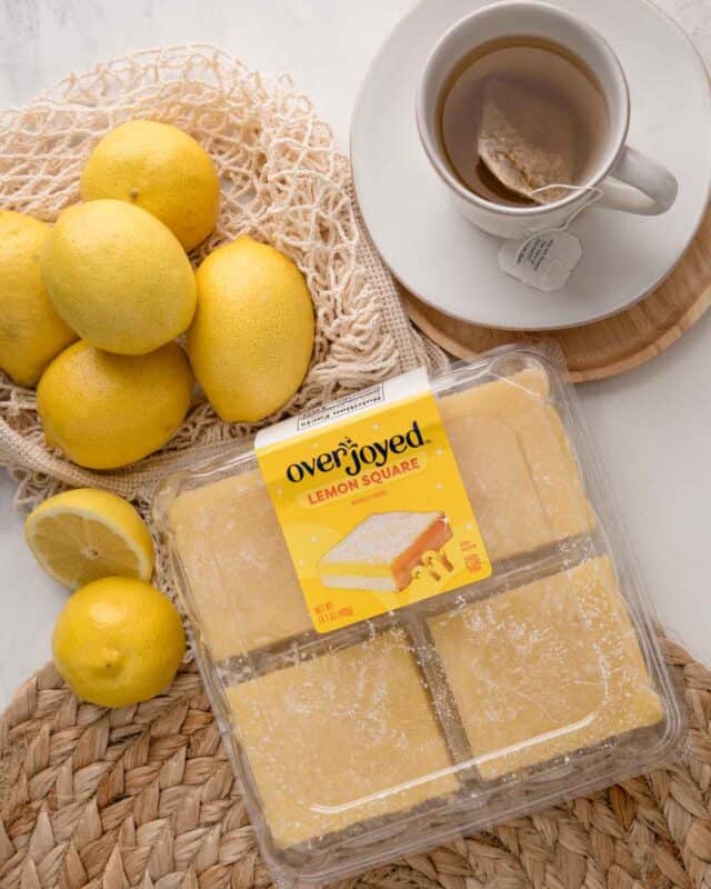 Lemon bars in packaging next to a pile of lemons and a cup of tea on a white marble countertop