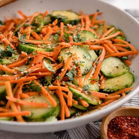 A salad with sliced cucumbers and carrots in a white bowl, garnished with sesame seeds and herbs.