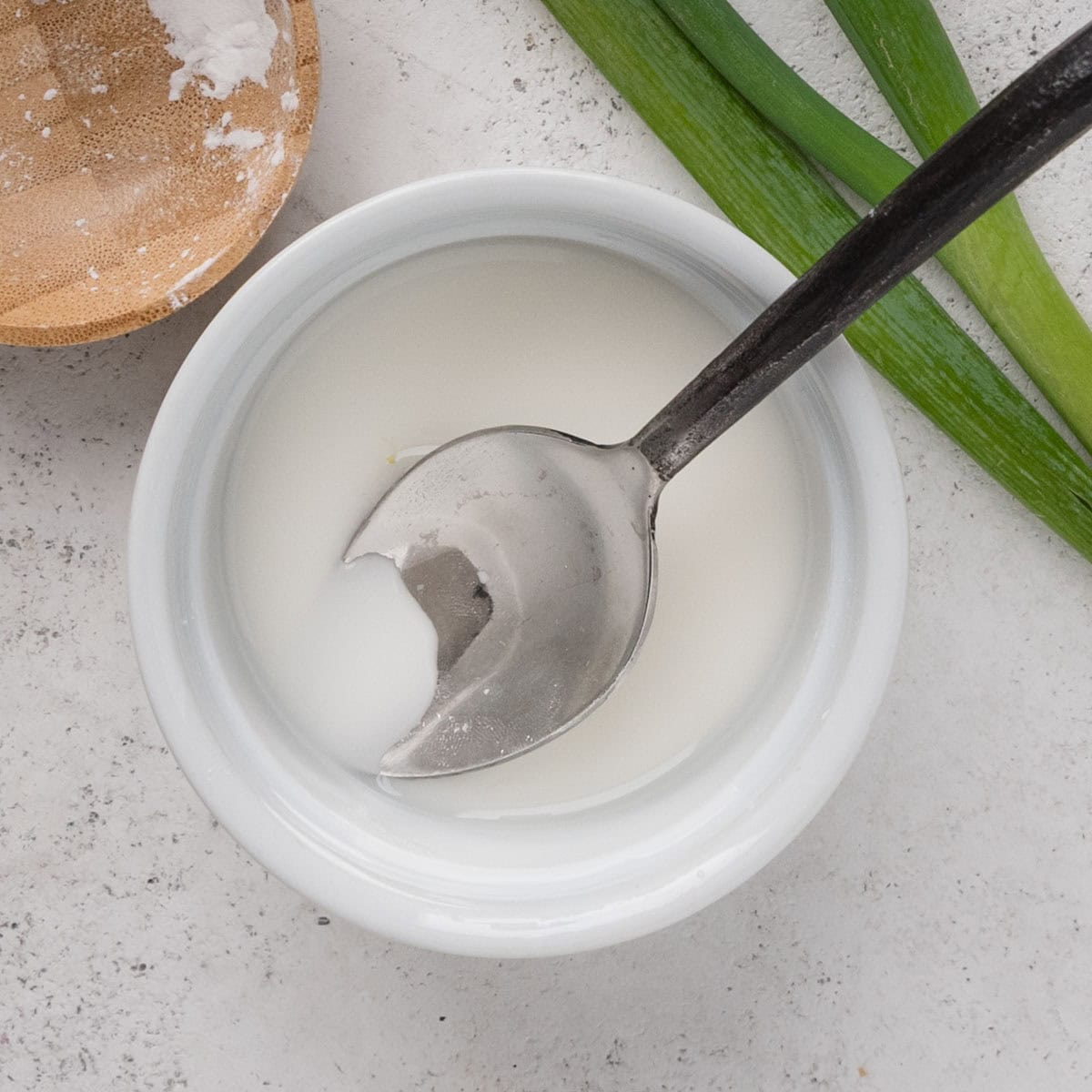 A metal spoon rests in a small white bowl of thick white liquid on a light surface, with green onions and a wooden bowl nearby.