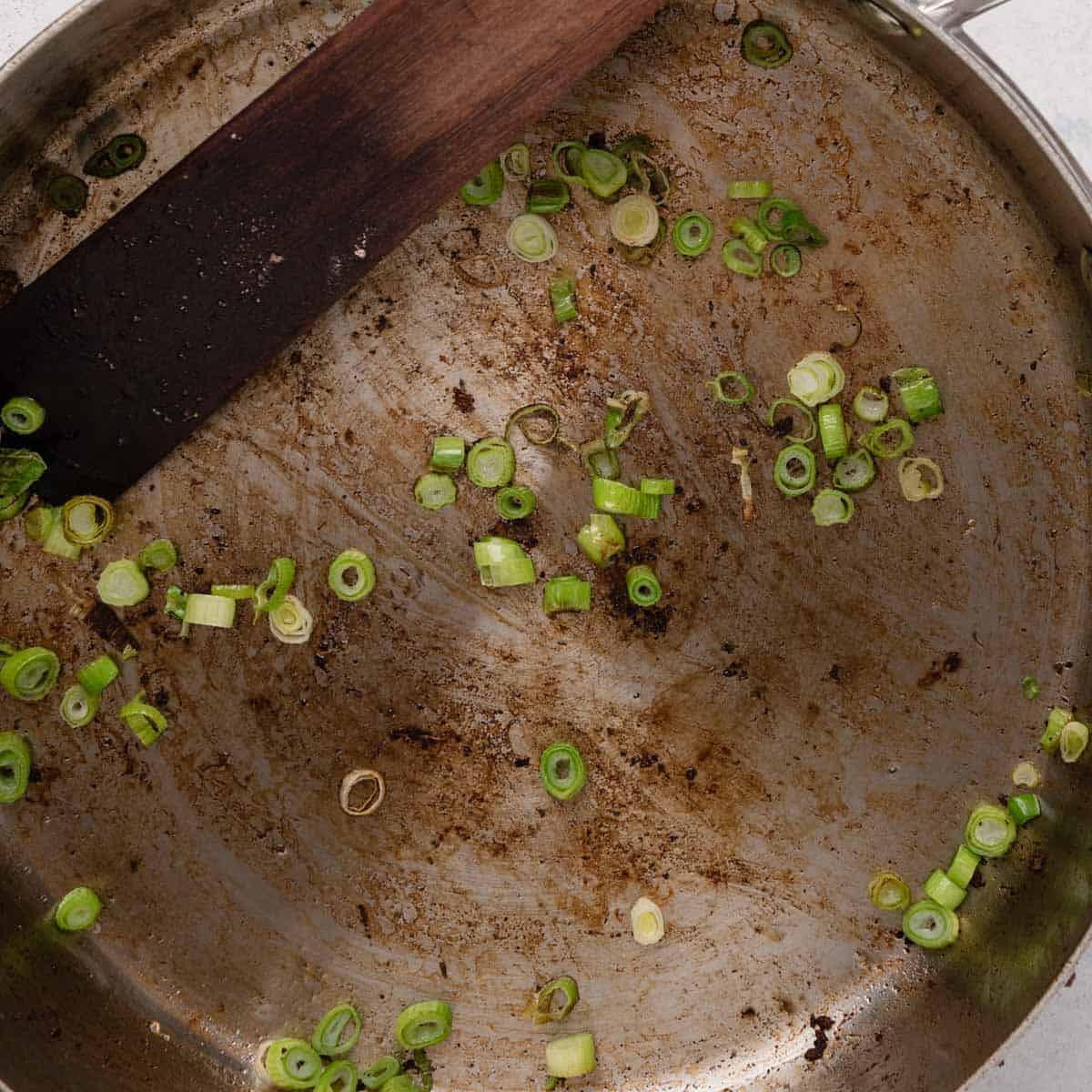A used pan with scattered chopped green onions and browned residue, alongside a dark wooden spatula.