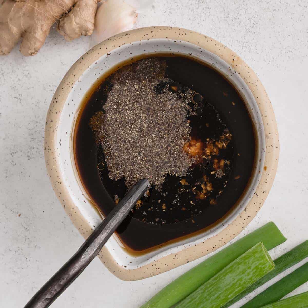 A bowl of dark soy sauce with a sprinkling of ground pepper and minced garlic, accompanied by a spoon. Ginger and green onions are nearby on a light surface.