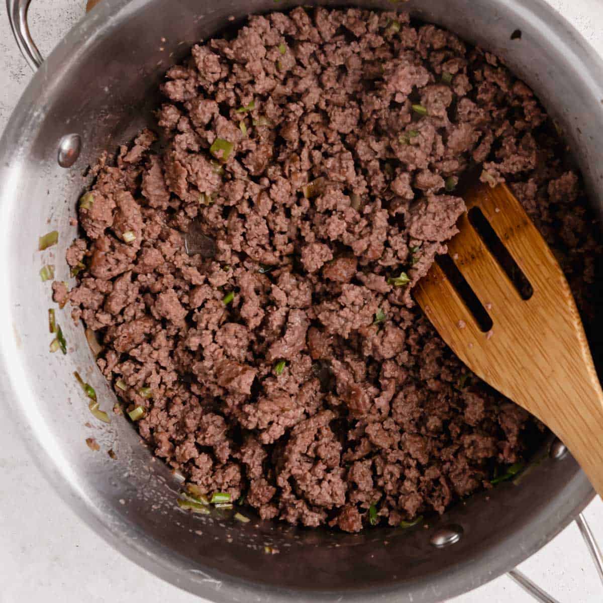 Ground meat cooking in a stainless steel pan with chopped green herbs, alongside a wooden spatula.