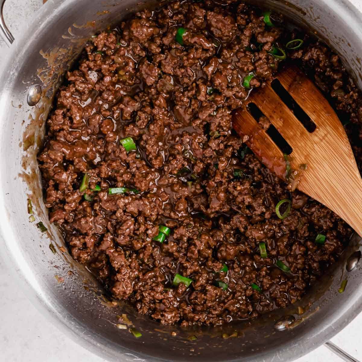 Cooked ground beef with chopped green onions in a stainless steel pan, stirred with a wooden spatula.