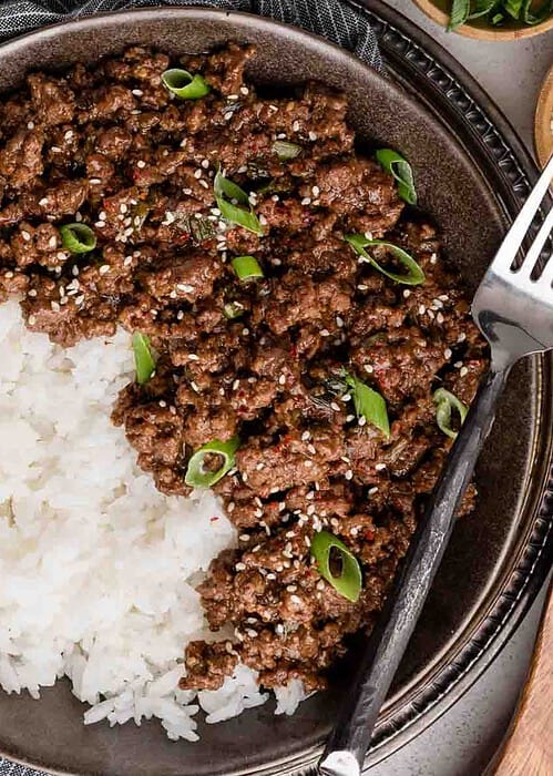 A bowl of white rice topped with ground beef garnished with sliced green onions and sesame seeds, accompanied by a fork.