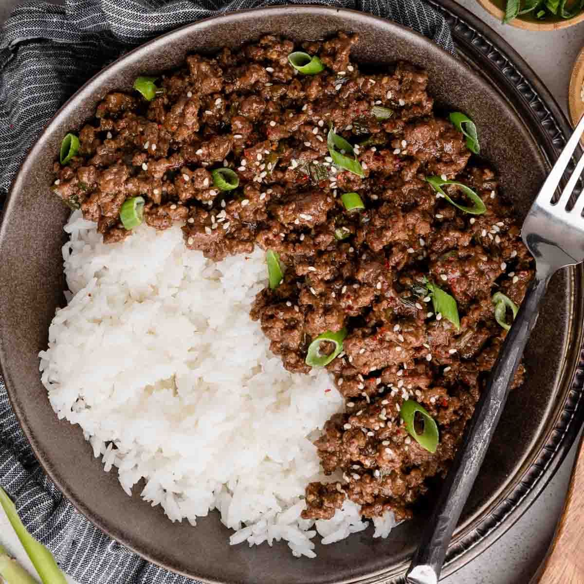 A bowl of white rice topped with Mongolian ground beef garnished with sliced green onions and sesame seeds, accompanied by a fork.