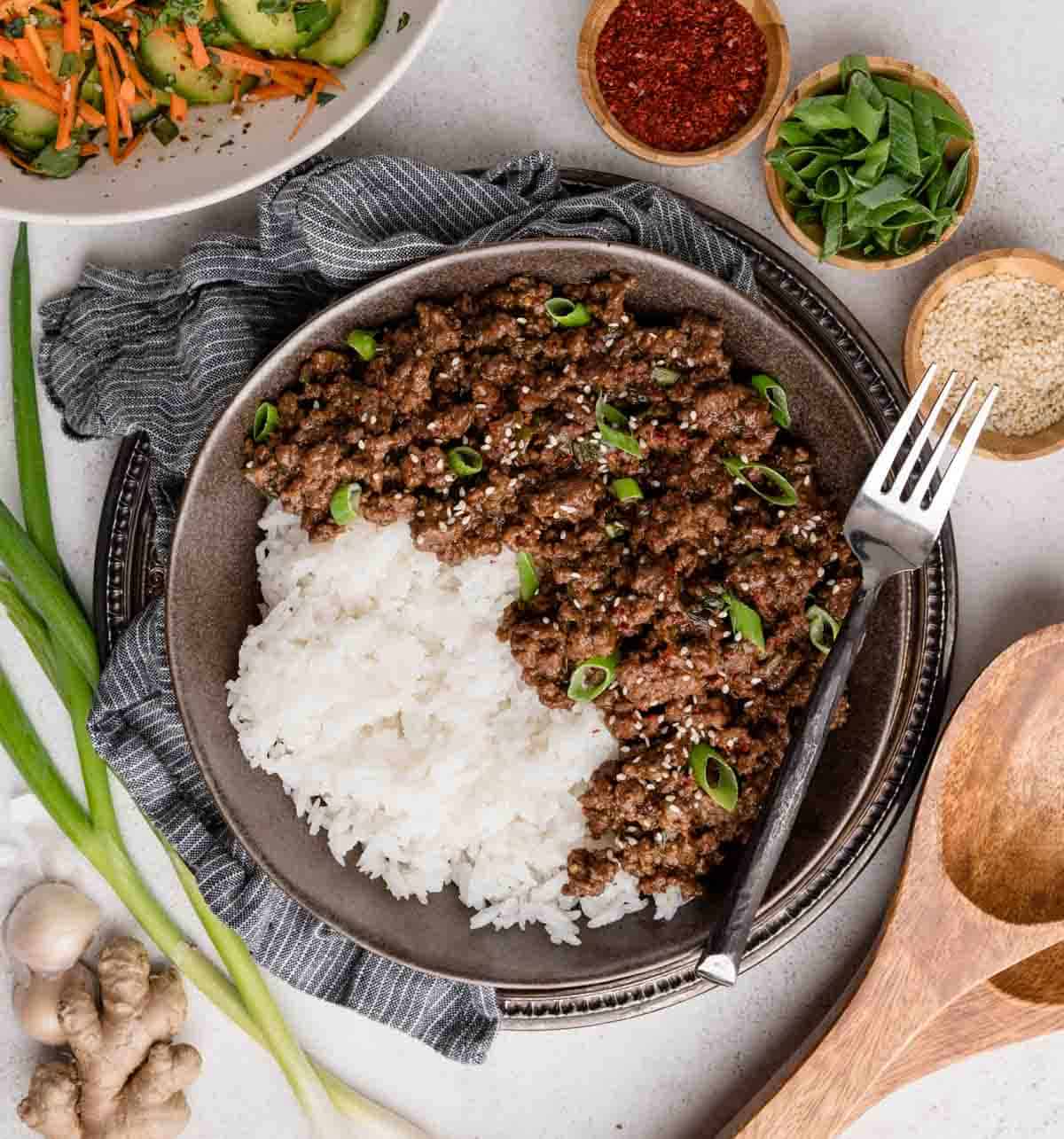 A bowl of white rice topped with minced beef, garnished with green onions, with various spices and vegetables surrounding it. A fork rests on the plate.