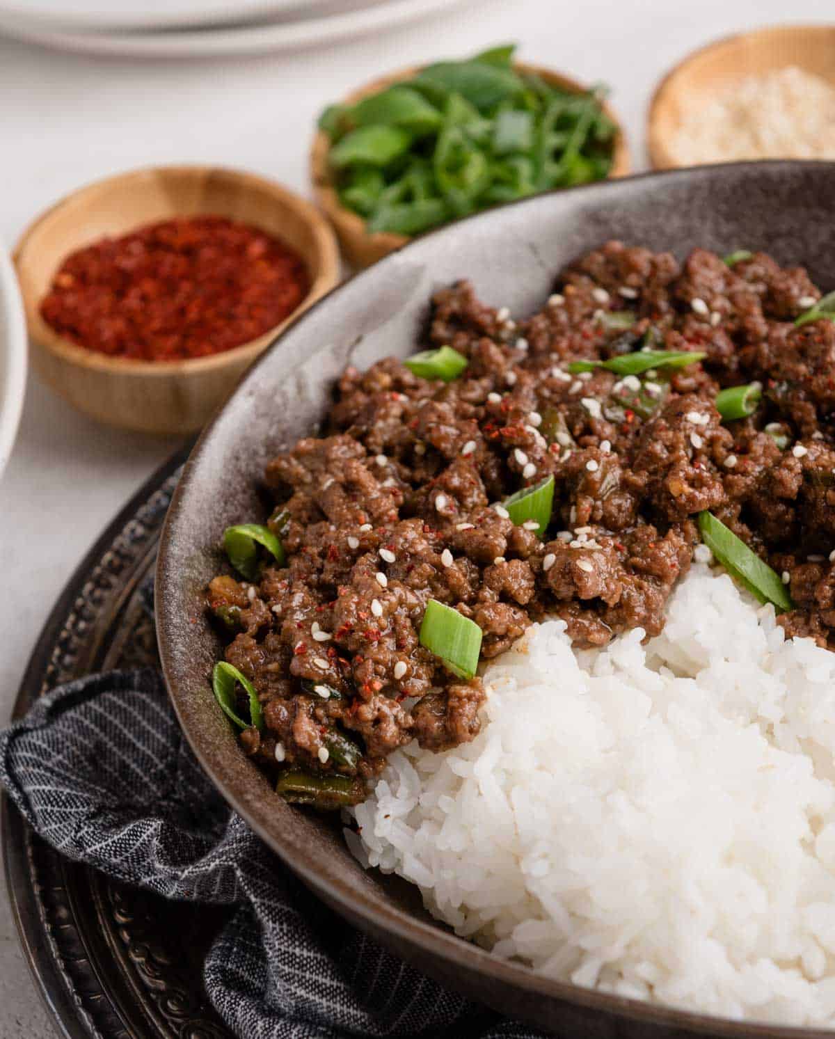 Bowl of white rice topped with seasoned ground beef, sprinkled with sesame seeds and green onions, surrounded by small bowls of red spice, green onions, and sesame seeds.