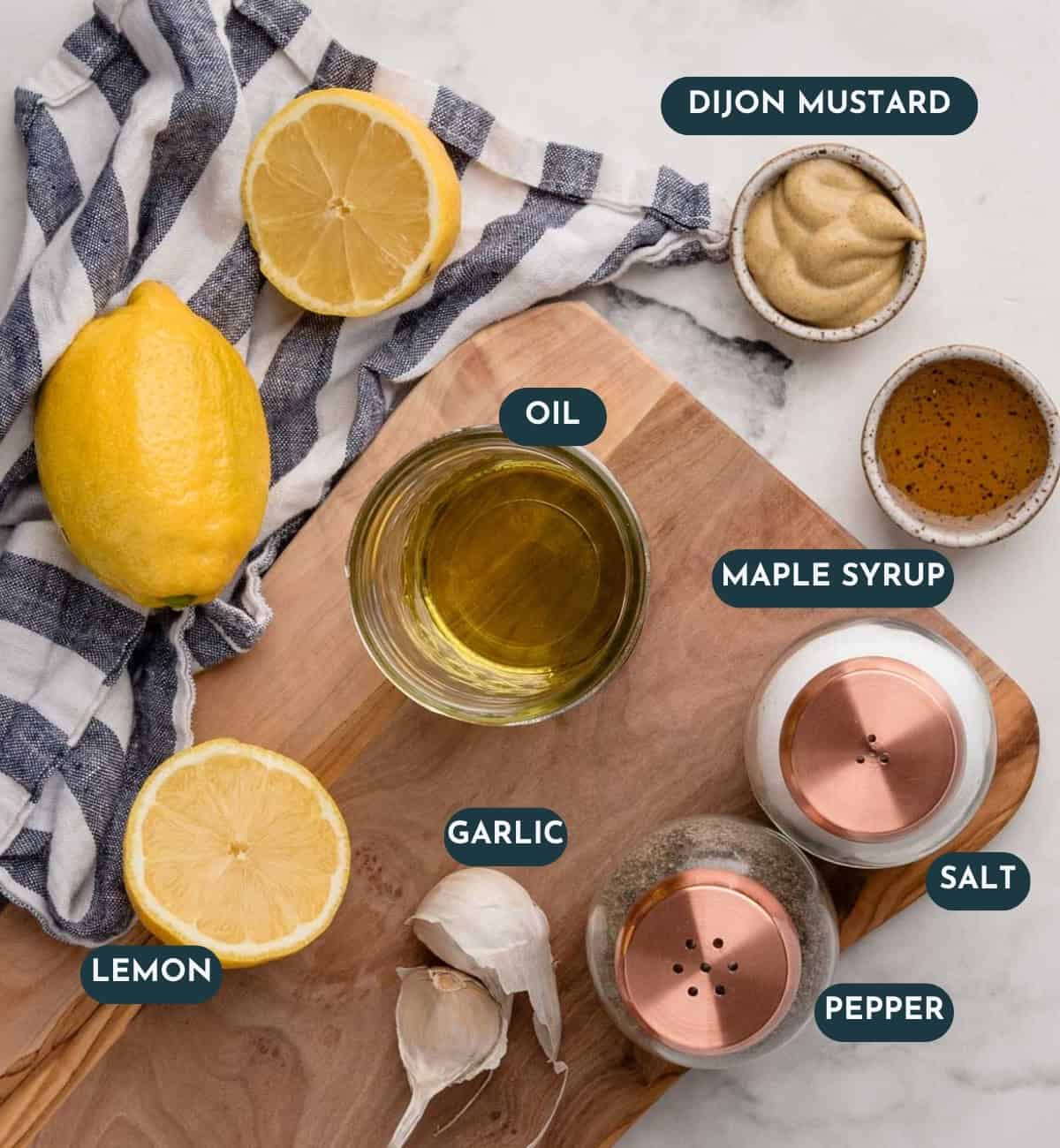 Ingredients for a Maple Dijon Dressing: lemon, oil, garlic, salt, pepper, Dijon mustard, and maple syrup arranged on a wooden board.
