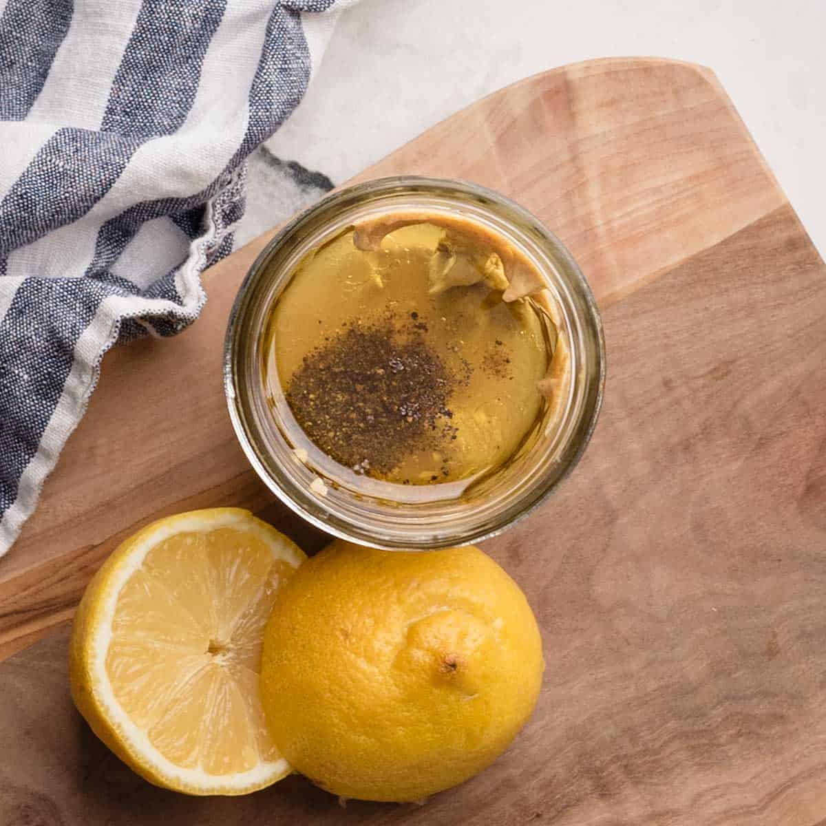 A glass jar with mustard, oil, and black pepper next to a whole lemon and a halved lemon on a wooden cutting board with a striped cloth nearby.