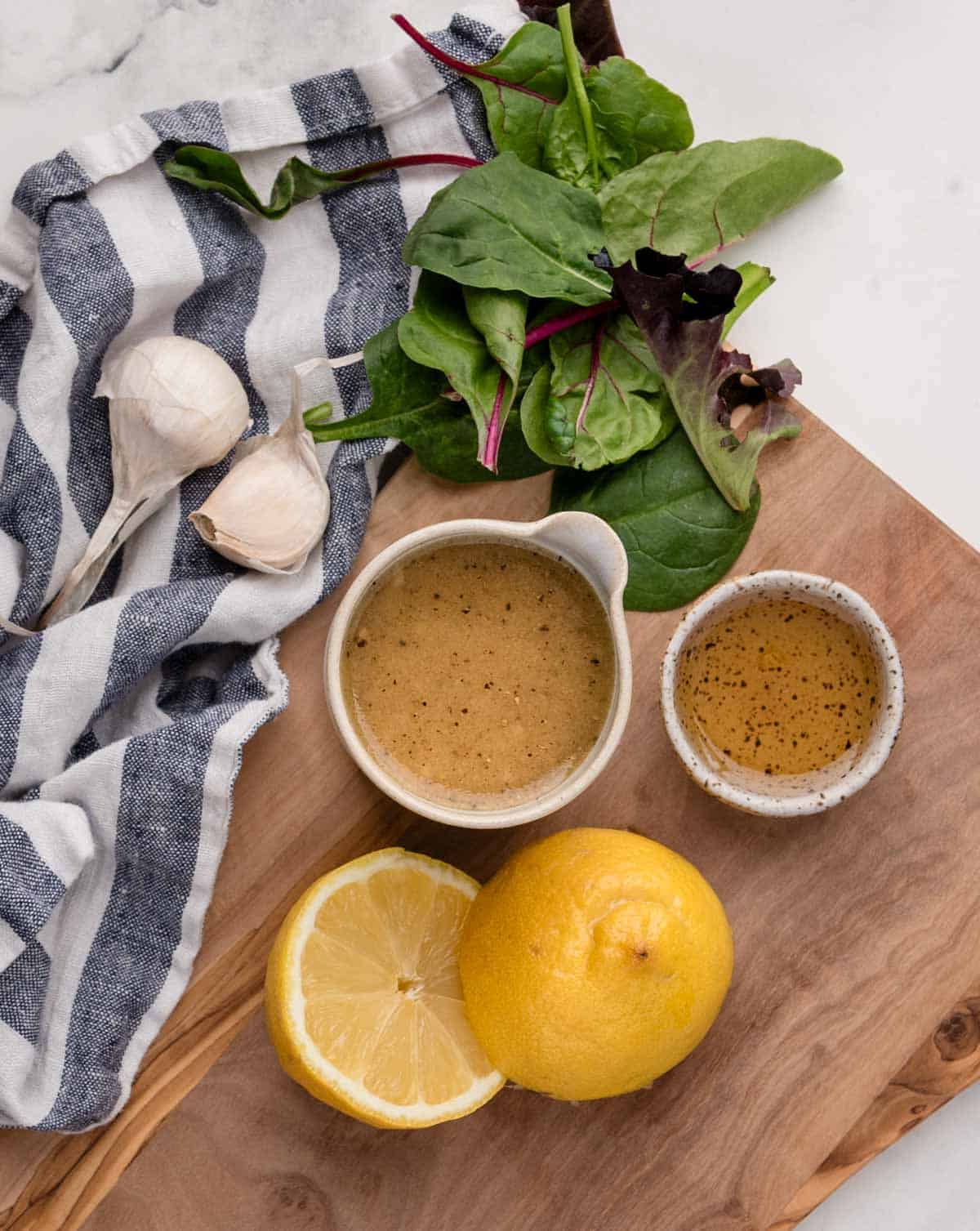 A wooden board with a halved lemon, mixed greens, garlic bulbs, a bowl and small cup of maple dijon vinaigrette, and a striped cloth.