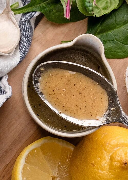 A spoon rests in a small bowl of maple dijon dressing, surrounded by spinach leaves, garlic cloves, a halved lemon, and a striped cloth on a wooden surface.