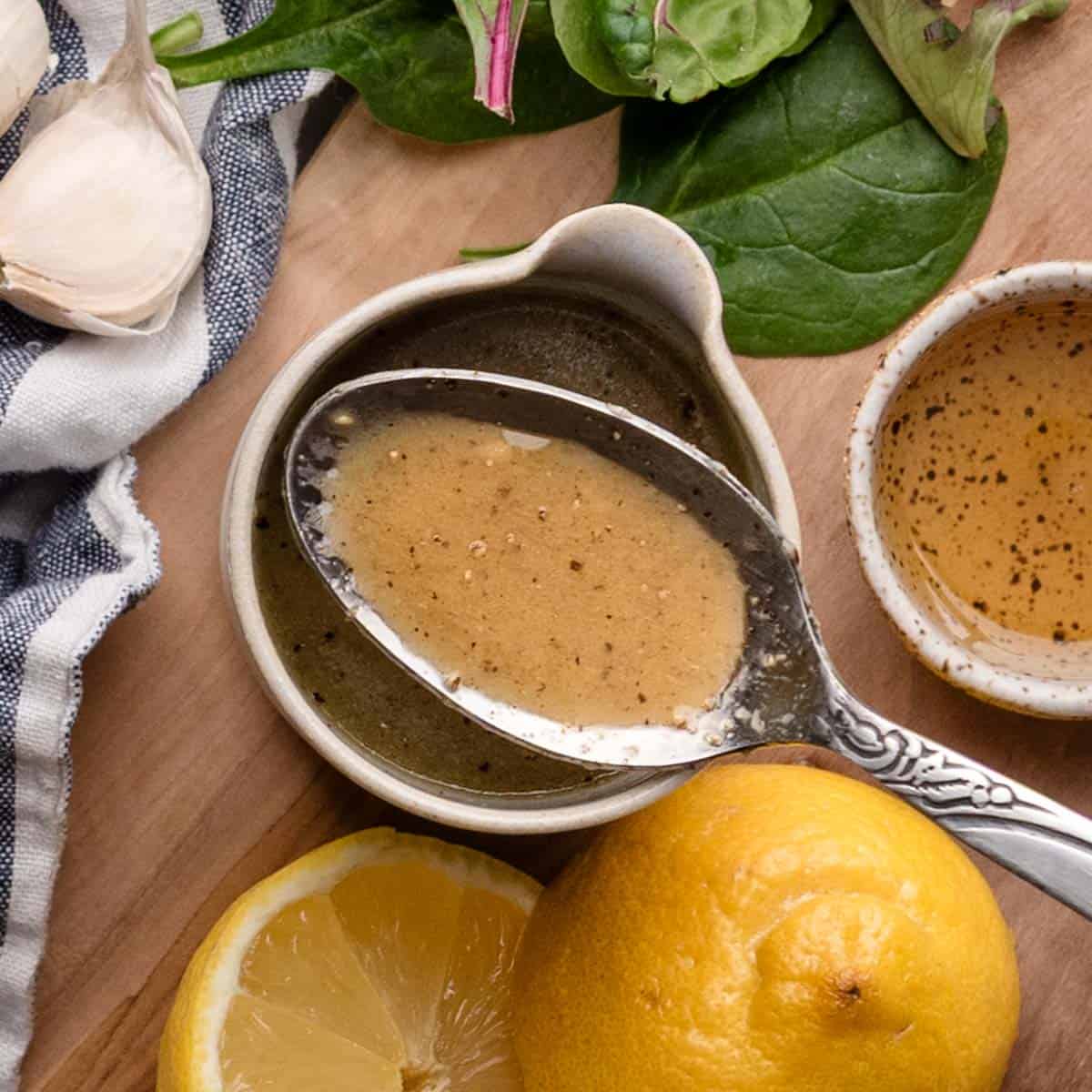 A spoon rests in a small bowl of maple dijon dressing, surrounded by spinach leaves, garlic cloves, a halved lemon, and a striped cloth on a wooden surface.