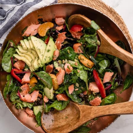 Smoked salmon salad with greens, avocado slices, salmon, red peppers, and oranges in a large bowl with wooden salad servers.