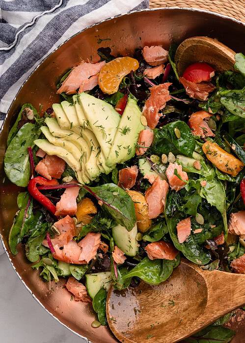 Smoked salmon salad with greens, avocado slices, salmon, red peppers, and oranges in a large bowl with wooden salad servers.
