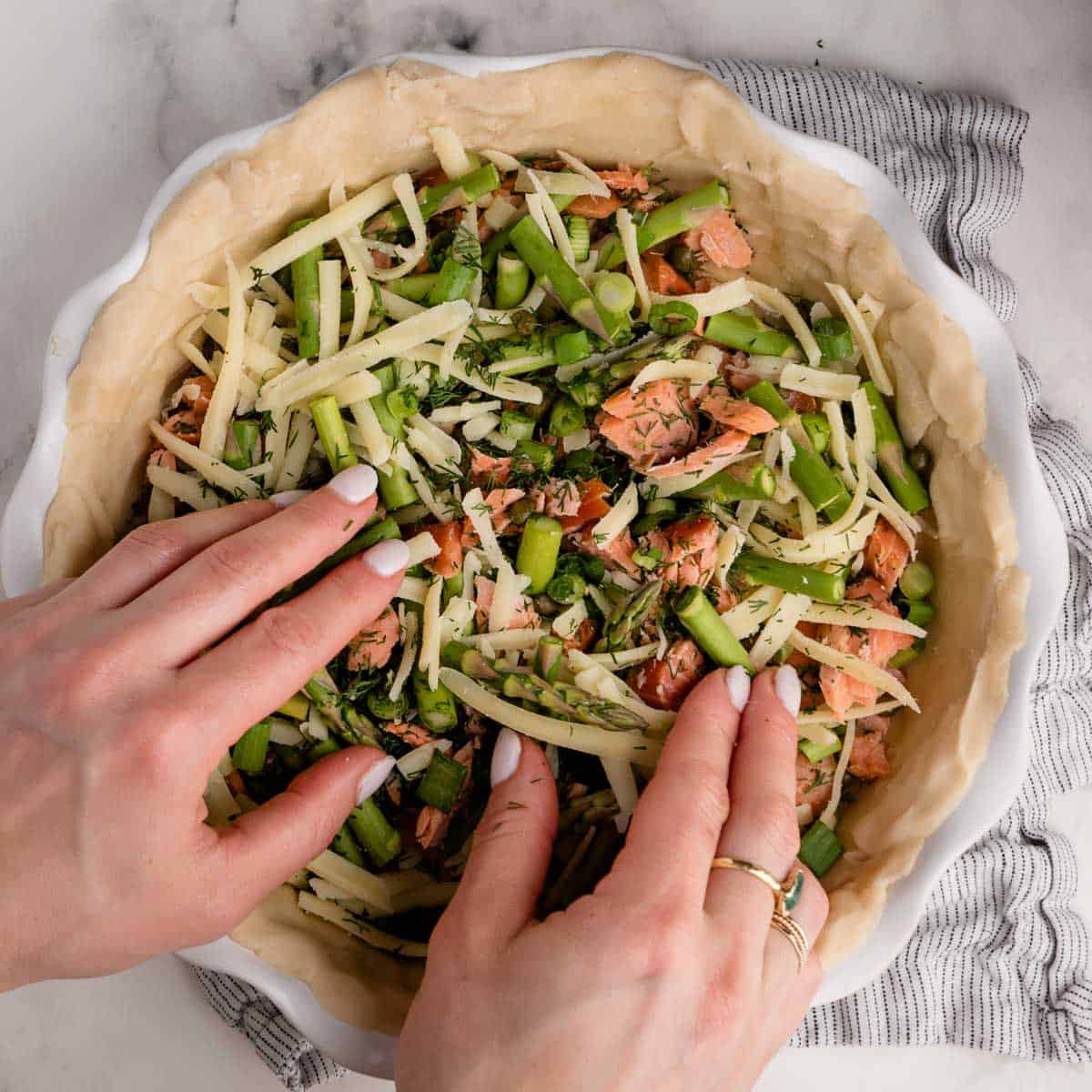 Hands arranging cheese, salmon, and green vegetables in a pie crust on a marble surface.
