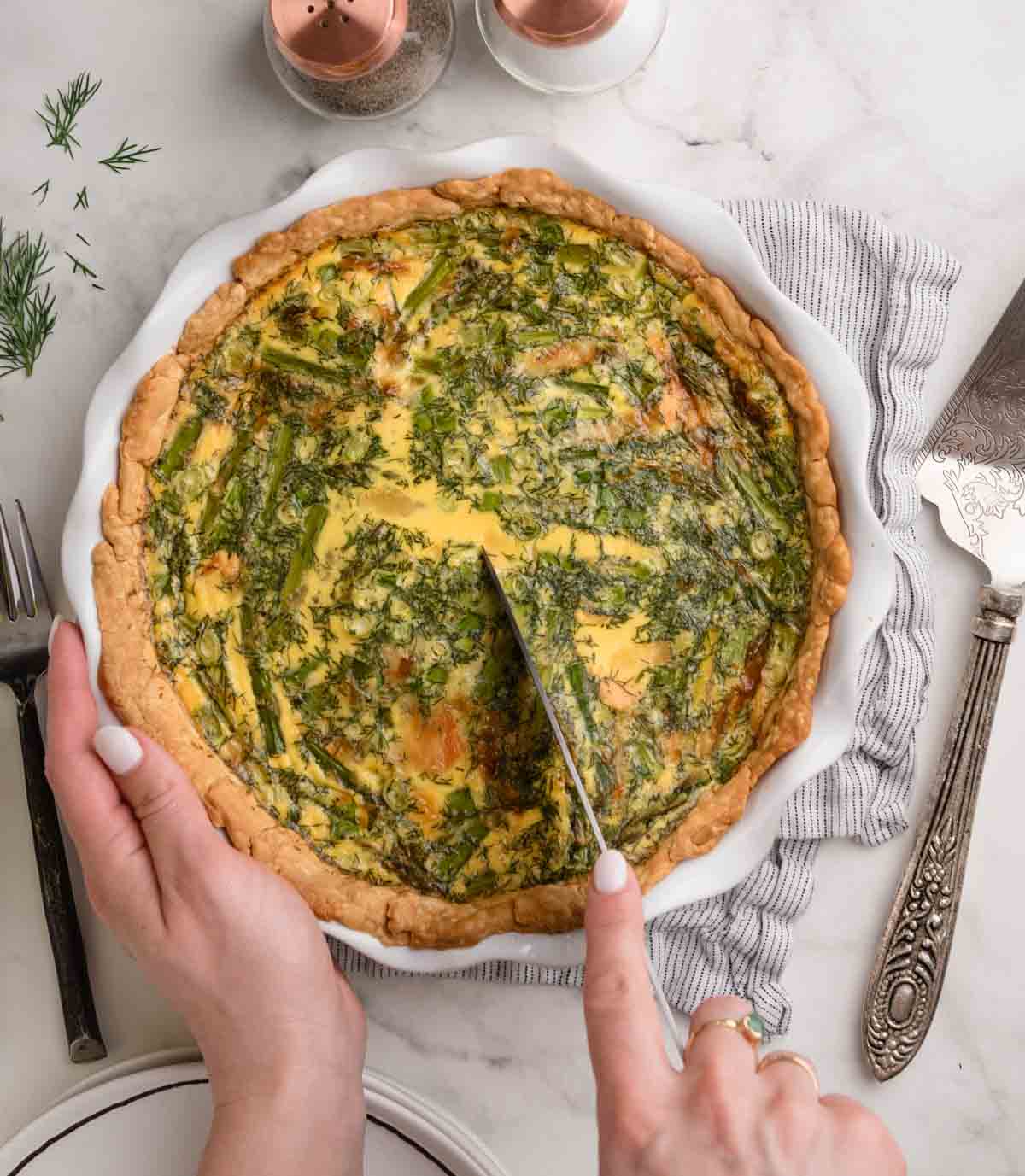 A person slices into a salmon, asparagus and dill quiche in a pie dish on a table set with utensils and a napkin.