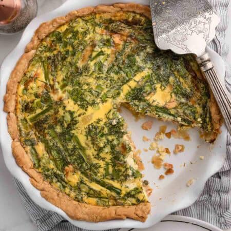 A baked smoked salmon quiche in a pie dish with one slice removed. Has lots of herbs and asparagus.