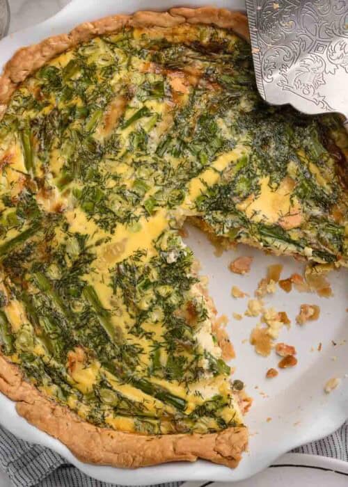 A baked smoked salmon quiche in a pie dish with one slice removed. Has lots of herbs and asparagus.