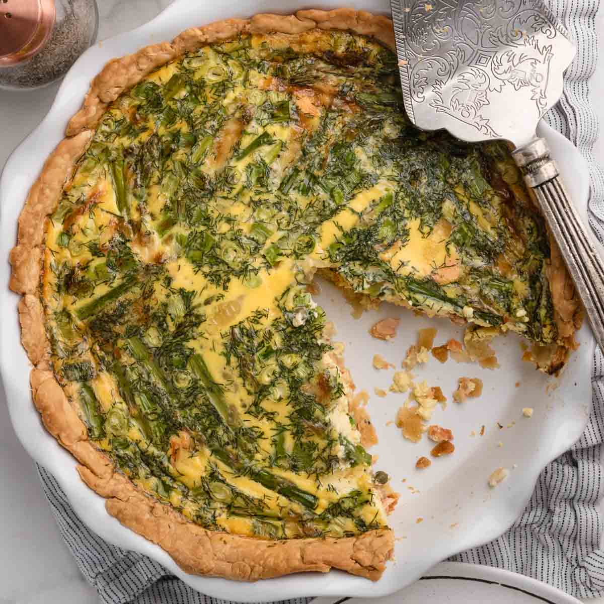 A smoked salmon quiche with dill and asparagus on top in a pie dish with one slice removed and a pie server resting on top.
