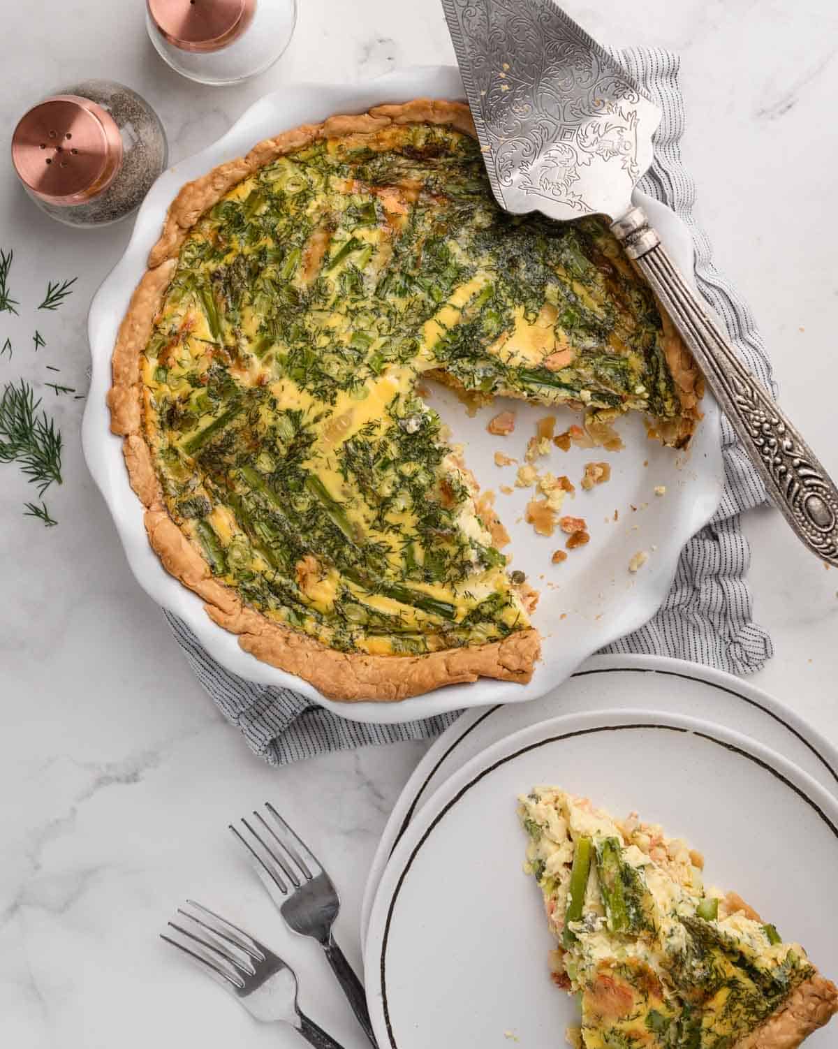 A vegetable quiche with a slice cut out, served on a plate beside forks and salt and pepper shakers.
