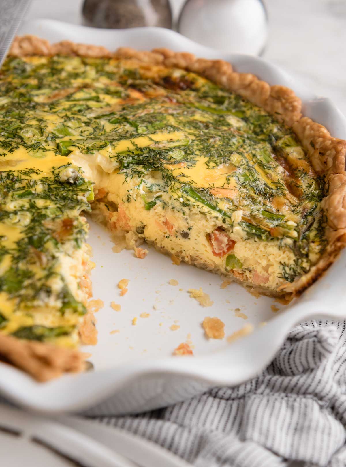 A baked smoked salmon quiche with herbs, vegetables, and a flaky crust, missing a large slice on a white dish.