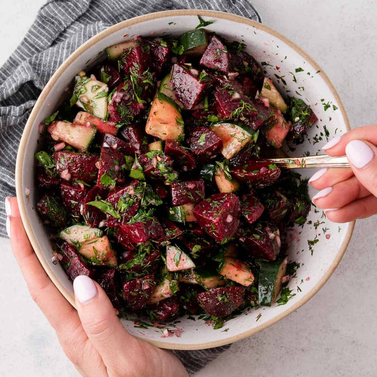 A bowl of beet and cucumber salad with herbs, held by two hands, one holding a fork.