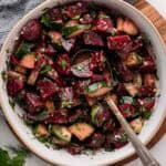 A bowl of beet and cucumber salad with herbs, mixed with a spoon.