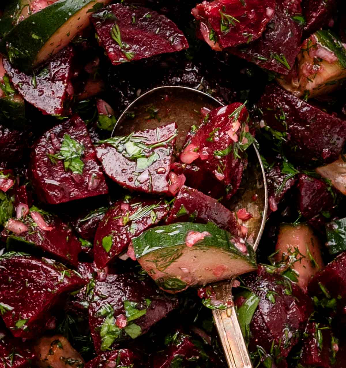 A close-up of beet and cucumber salad with herbs and vinaigrette, served with a silver spoon.
