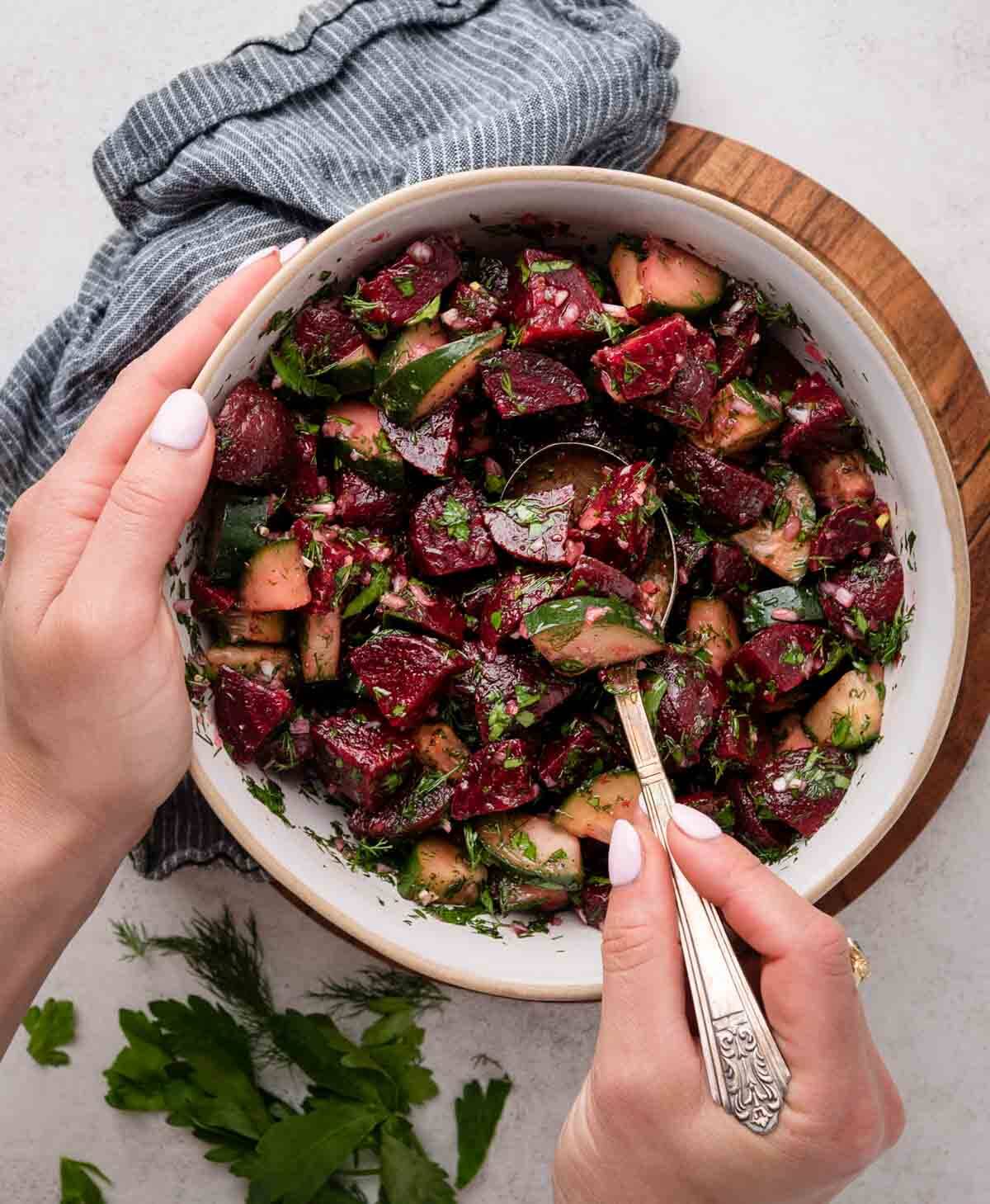A bowl of beet and cucumber salad with herbs, being mixed by a person holding a spoon.