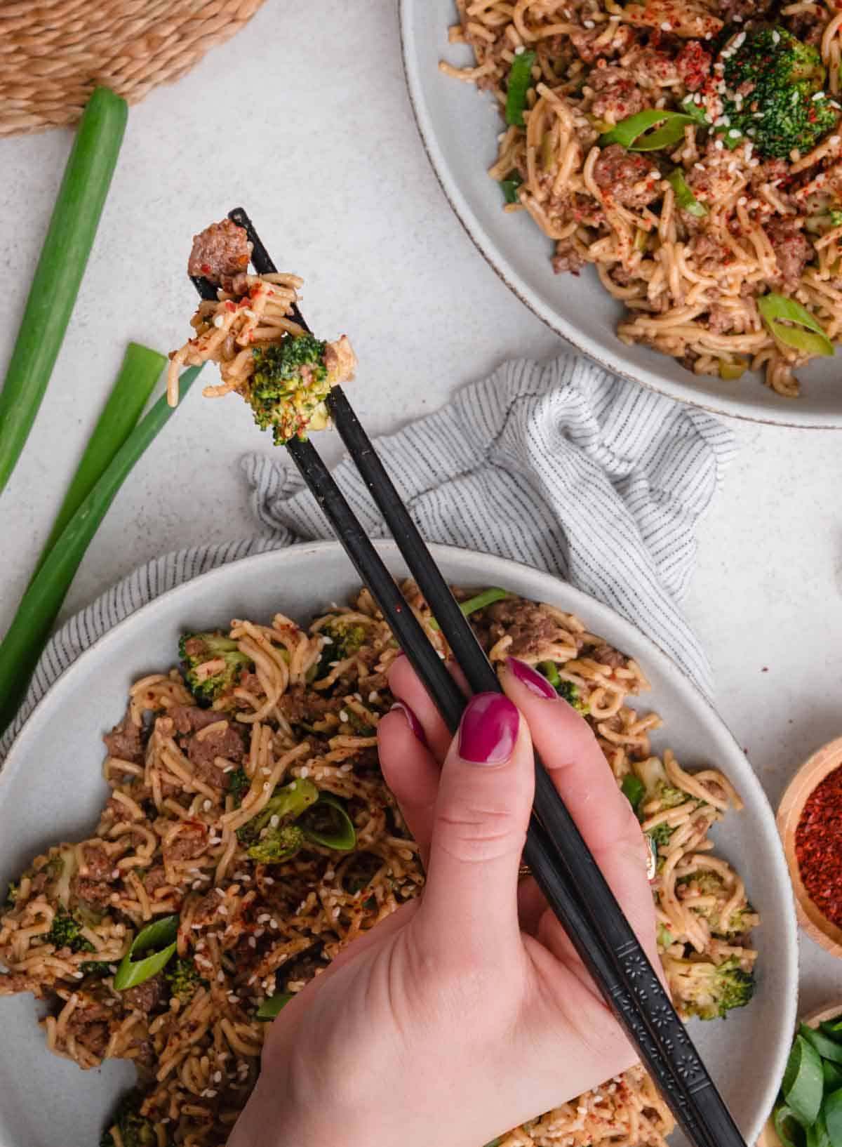 A hand with pink nails holds chopsticks picking up noodles and vegetables from a bowl of stir-fry.