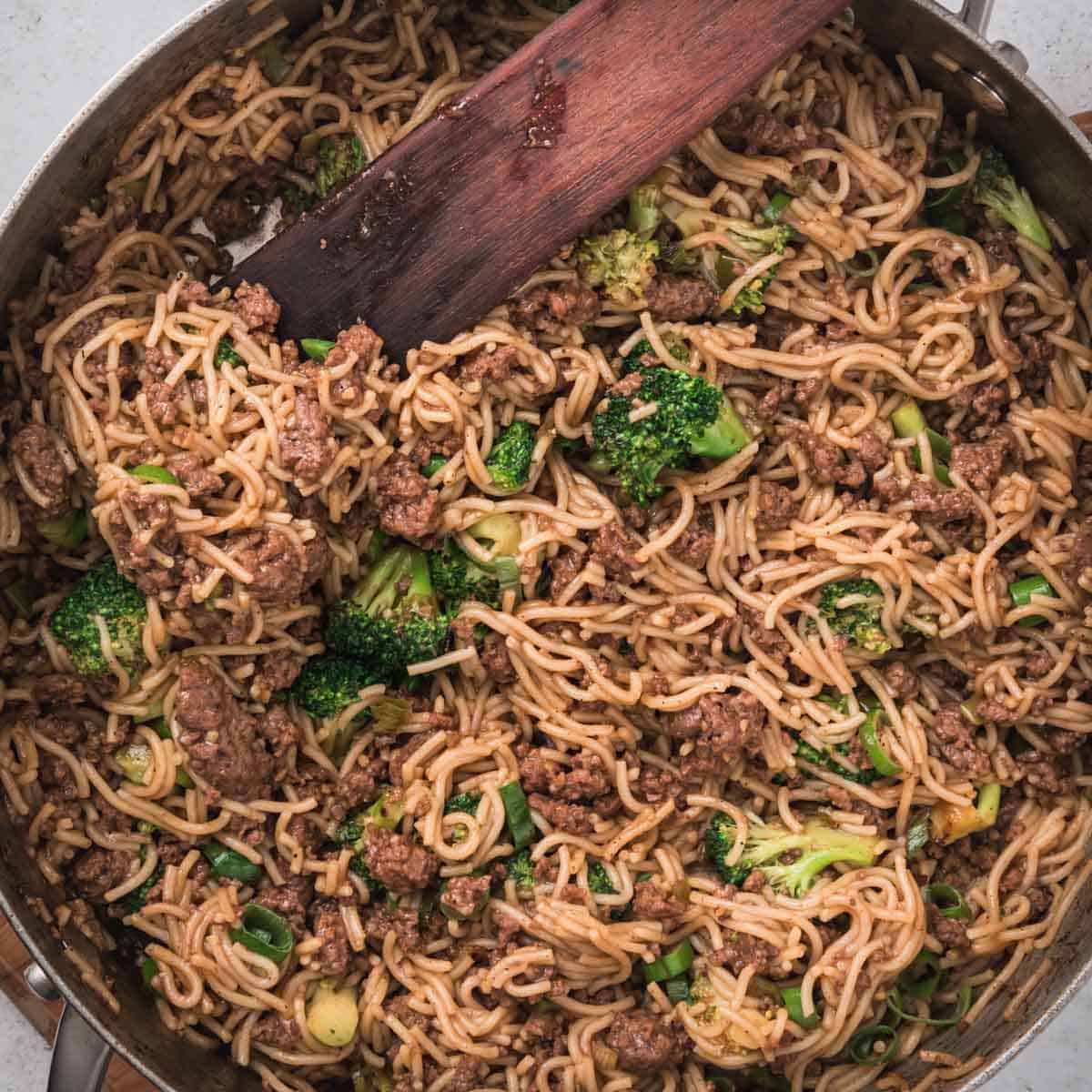 Pan of stir-fried noodles with ground beef and broccoli, mixed with a wooden spatula.