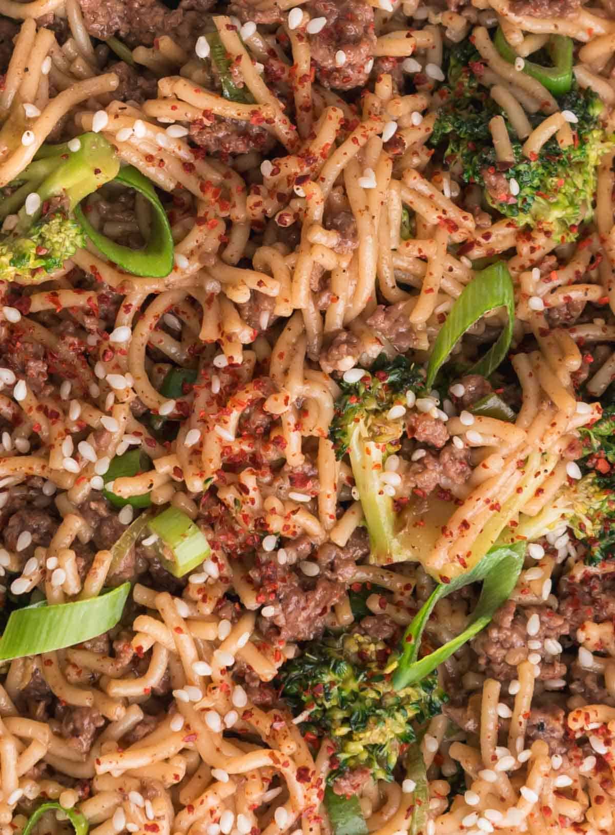Close-up of noodle stir fry with ground beef, broccoli, green onions, sesame seeds, and red pepper flakes.