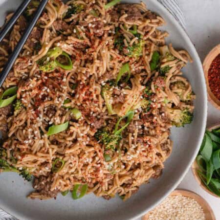 Mongolian Ground Beef Noodles with broccoli, green onions, sesame seeds, and red pepper flakes on a plate with chopsticks.