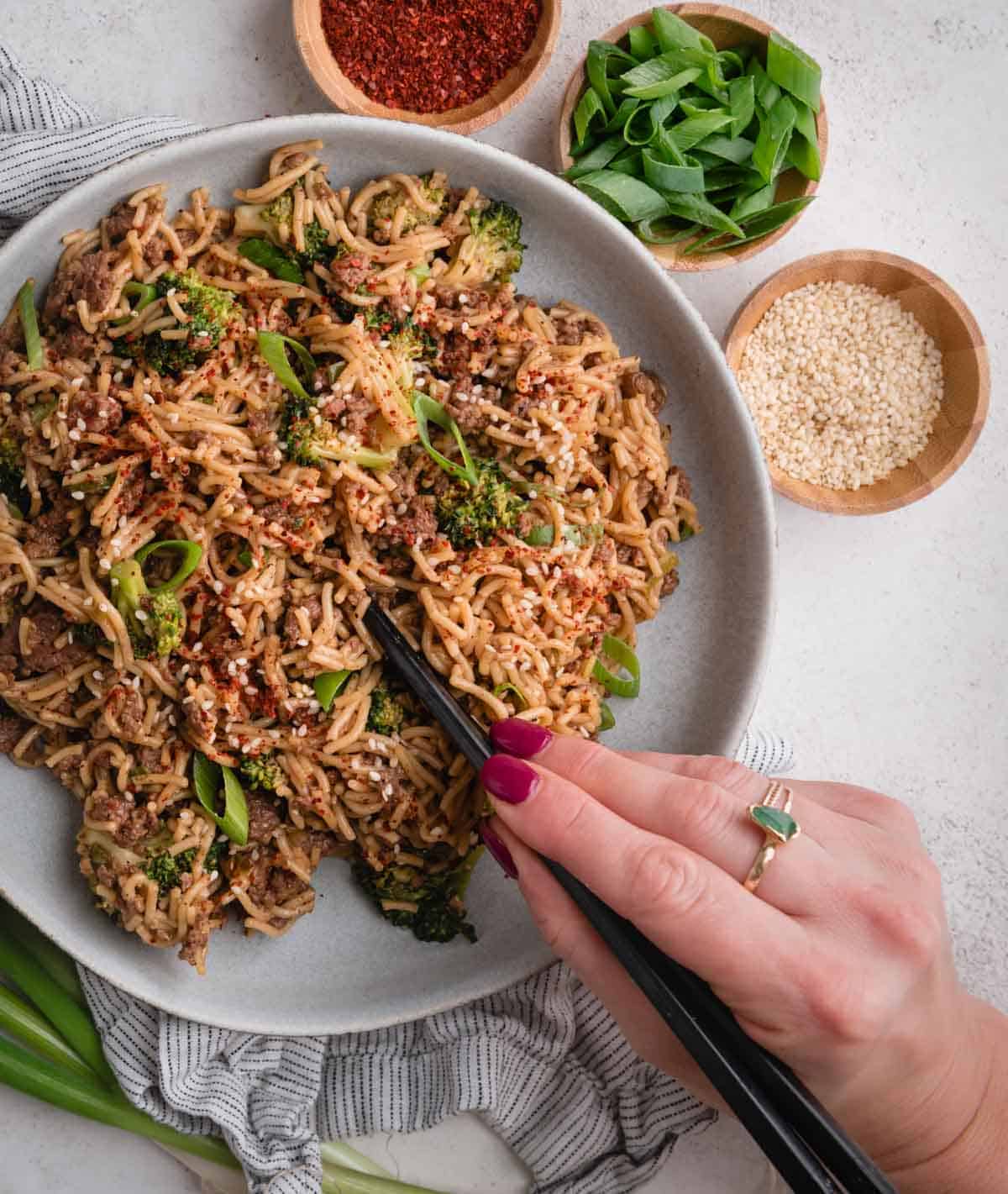 A hand using chopsticks to pick up beef and broccoli noodles, with green onions, sesame seeds, and spices nearby.
