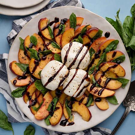Peach caprese salad with sliced peaches, mozzarella, and basil on a plate, drizzled with balsamic glaze, on a blue background.