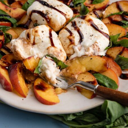 Peach caprese salad with sliced peaches, burrata cheese, fresh basil, and balsamic glaze on a plate, with a fork cutting in.