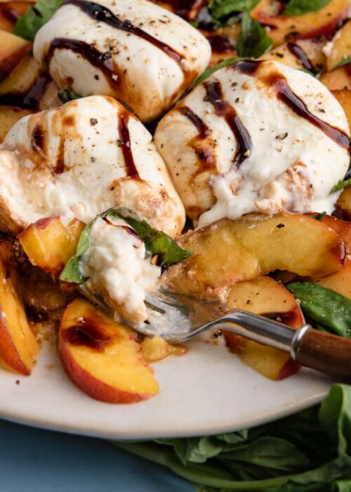 Peach caprese salad with sliced peaches, burrata cheese, fresh basil, and balsamic glaze on a plate, with a fork cutting in.