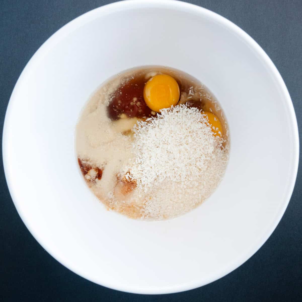 A white bowl with eggs, shredded coconut, and various wet ingredients ready to be mixed.