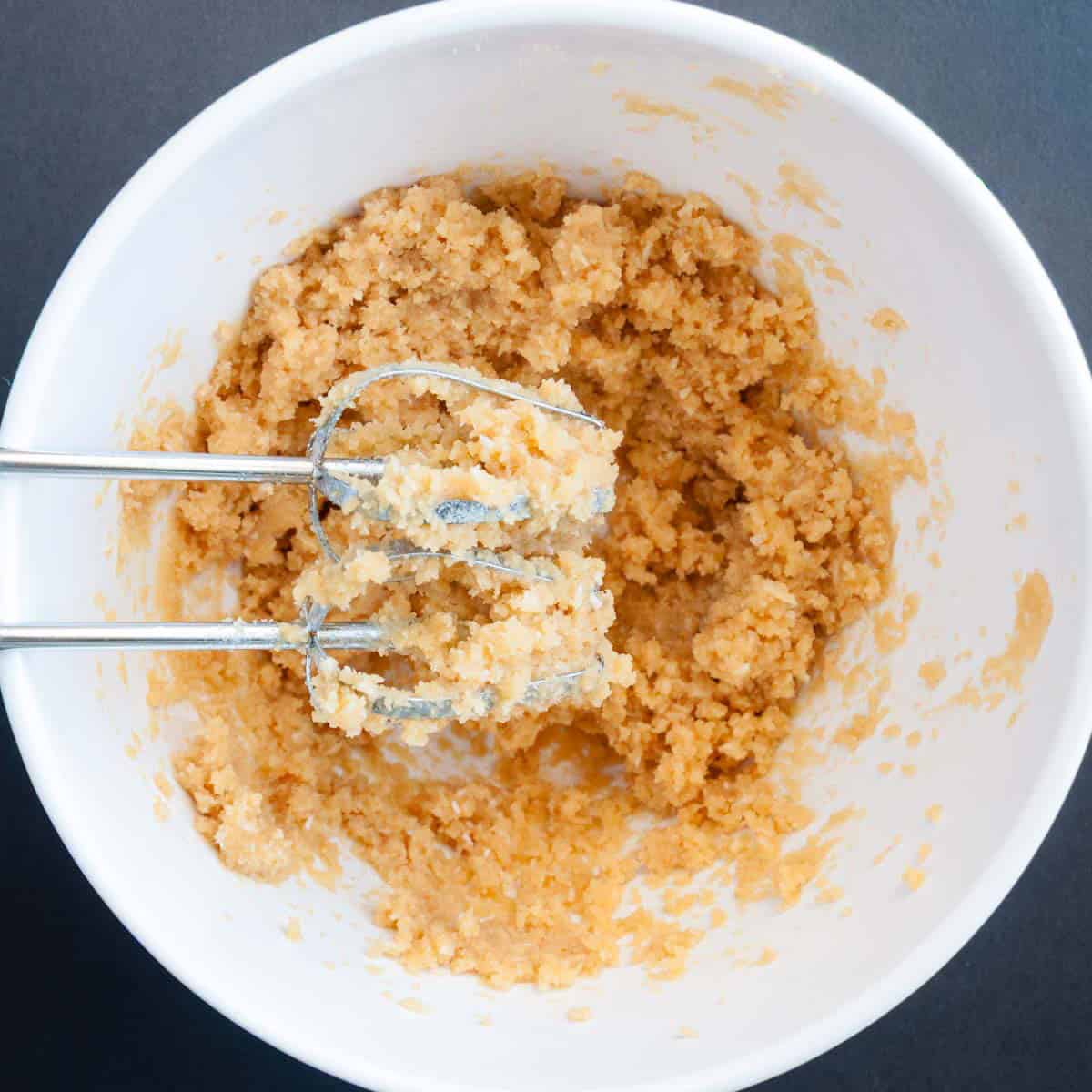 A white bowl with cookie dough being mixed by an electric hand mixer on a dark surface.