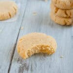 A bitten sugar cookie on a wooden surface with more cookies stacked in the background.