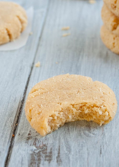A bitten sugar cookie on a wooden surface with more cookies stacked in the background.