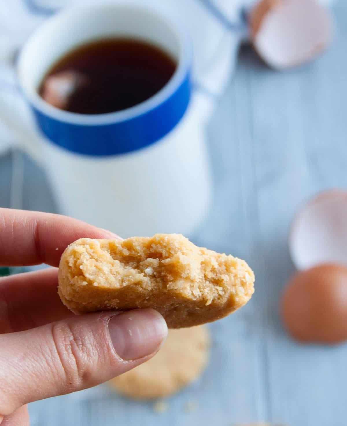 A hand holding a bitten coconut flour cookie with a cup of tea and eggshells in the background.