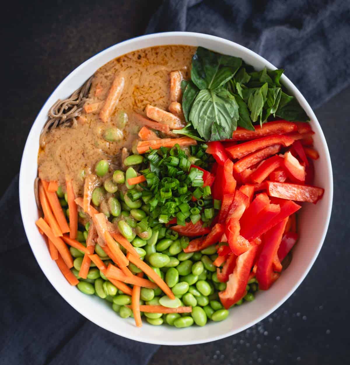 Bowl with sliced red peppers, carrots, edamame, green onions, basil, and a creamy sauce on noodles.
