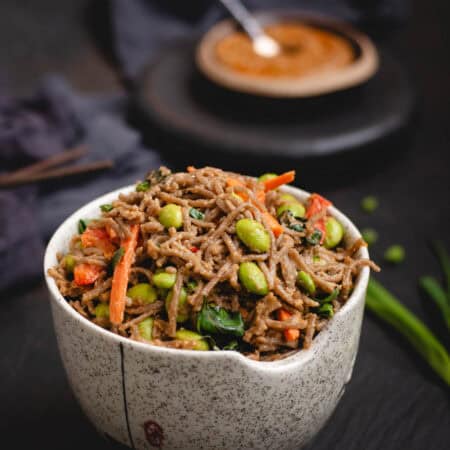 Bowl of soba noodles in peanut sauce with edamame, carrots, and Thai basil, with more peanut sauce in a dish in the background.