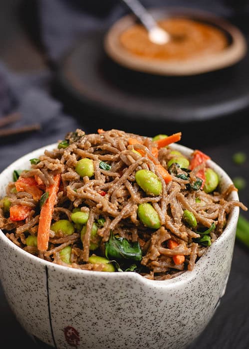 Bowl of soba noodles in peanut sauce with edamame, carrots, and Thai basil, with more peanut sauce in a dish in the background.