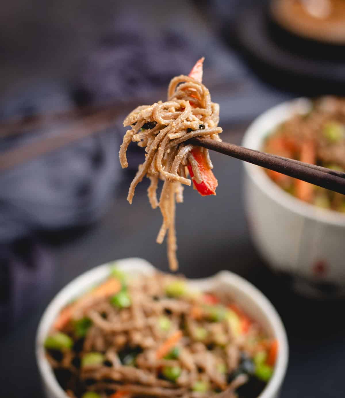 Chopsticks holding up a bite of soba noodles tossed in peanut sauce with vegetables above a bowl of noodles.