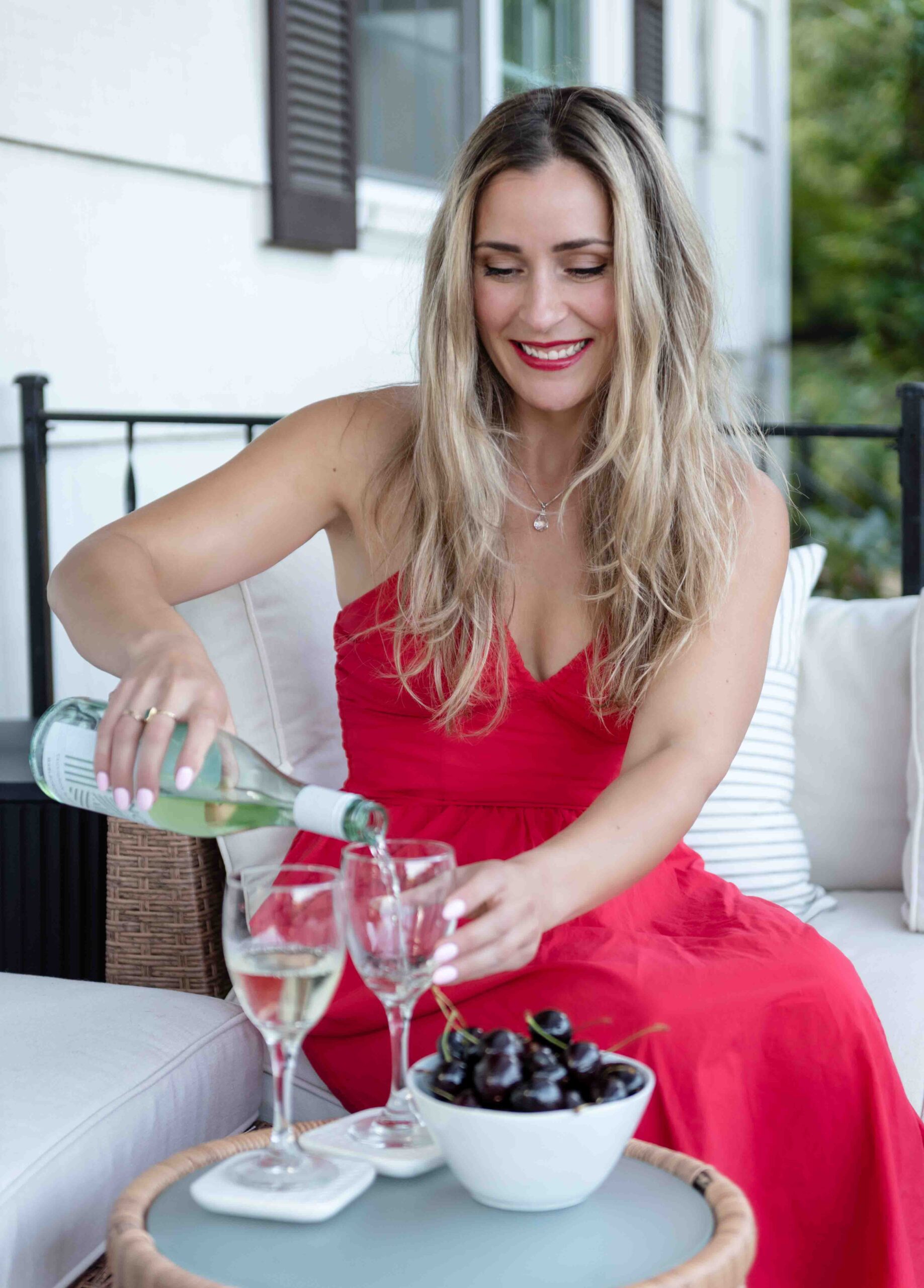 Woman in a red dress pours white wine into glasses on a patio table with a bowl of cherries.