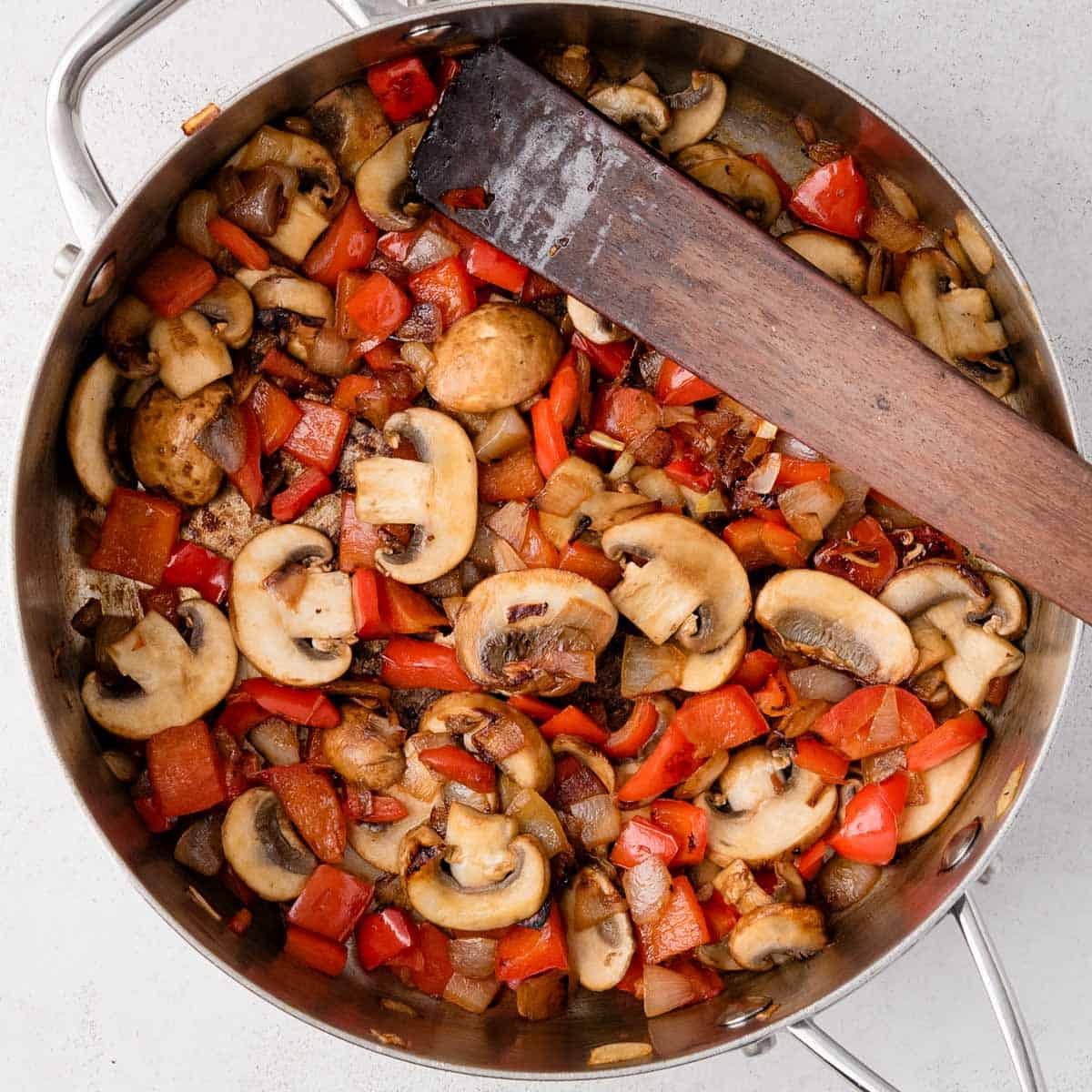 Sliced mushrooms, red bell peppers, and onions being sautéed in a pan with a wooden spatula.