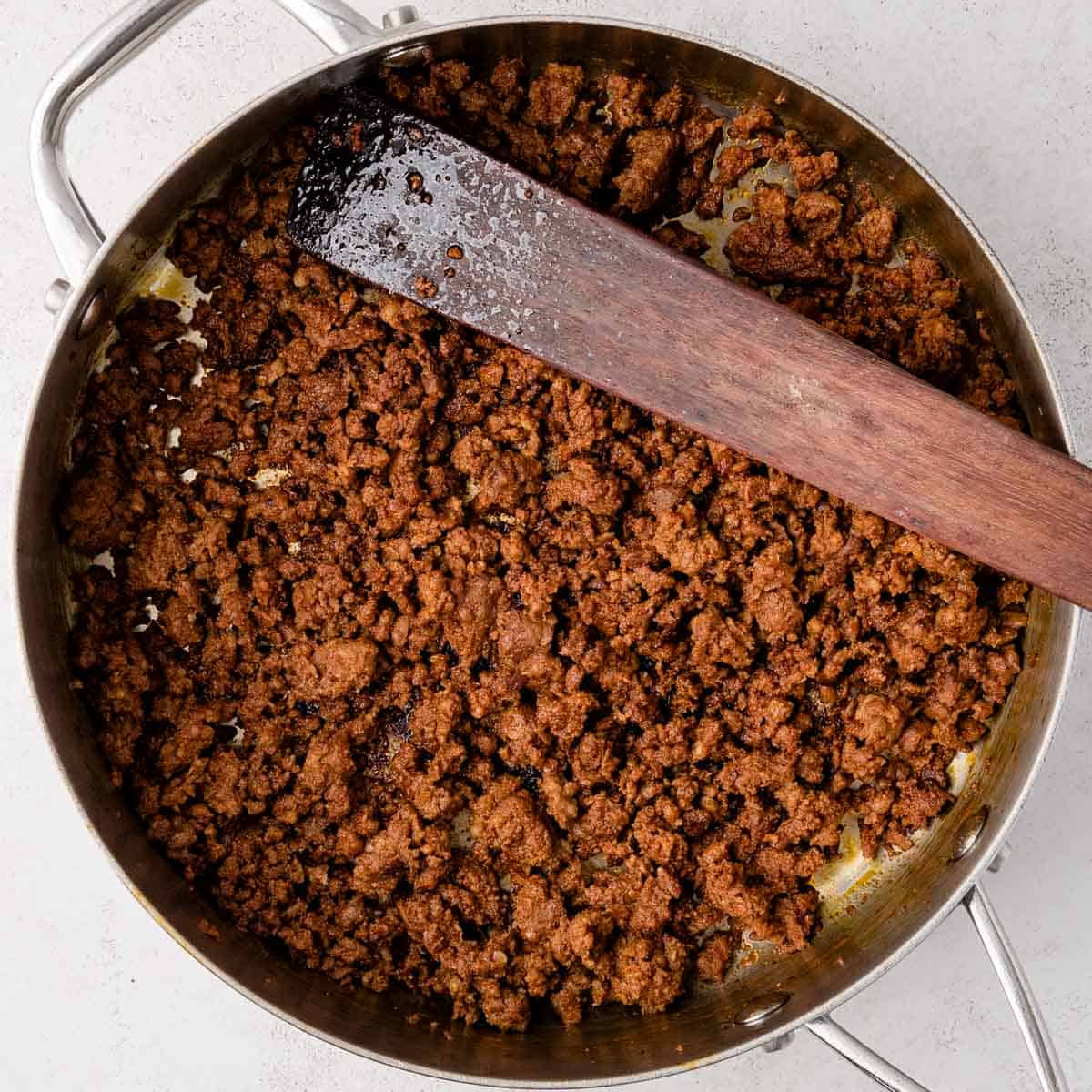 Cooked ground beef with seasoning in a stainless steel skillet, stirred with a wooden spatula.